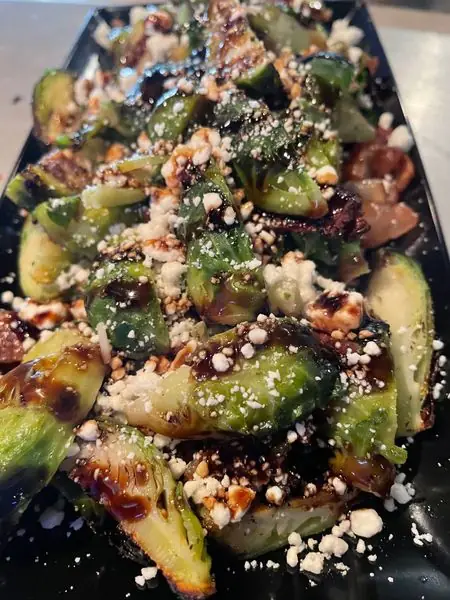 Wood fired brussel sprouts on a black plate, charred at the edges, finished with balsamic glaze and crumbled cheese