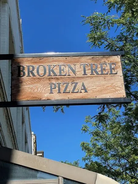 The Broken Tree Pizza hanging sign carved into wood outside the storefront in downtown Neenah, against a blue sky