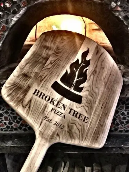 Broken Tree Pizza's own branded pizza peel, engraved Broken Tree Pizza Est. 2015, held up to the lit wood-fired oven