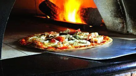 A pizza going into the lit wood-fired oven on a metal peel