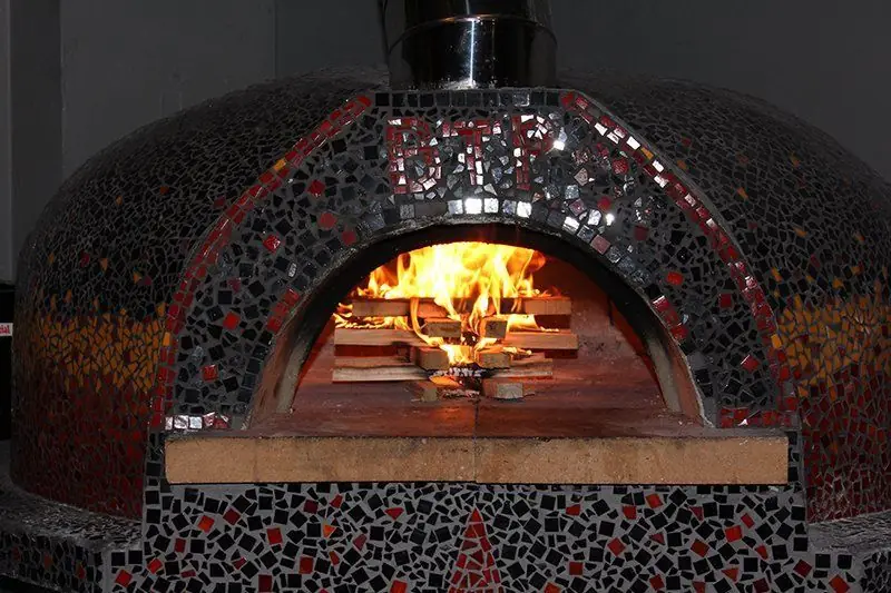 The wood-fired oven at Broken Tree Pizza, a mosaic-tiled dome with BTP set into the arch in mirror tile and a live fire burning on the stone floor