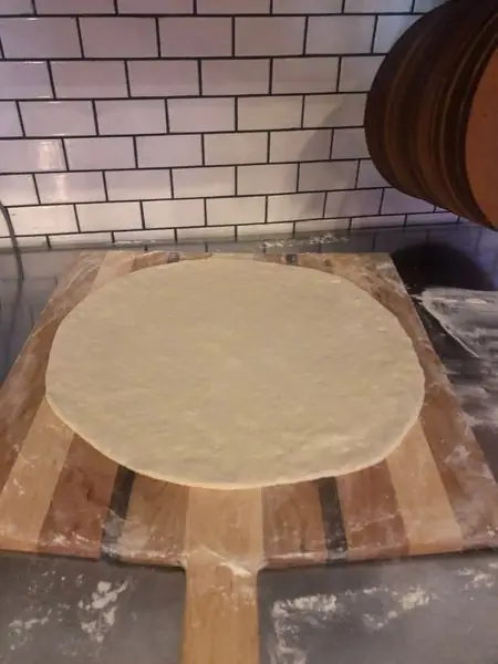 Hand-stretched raw pizza dough resting on a wooden peel against subway tile